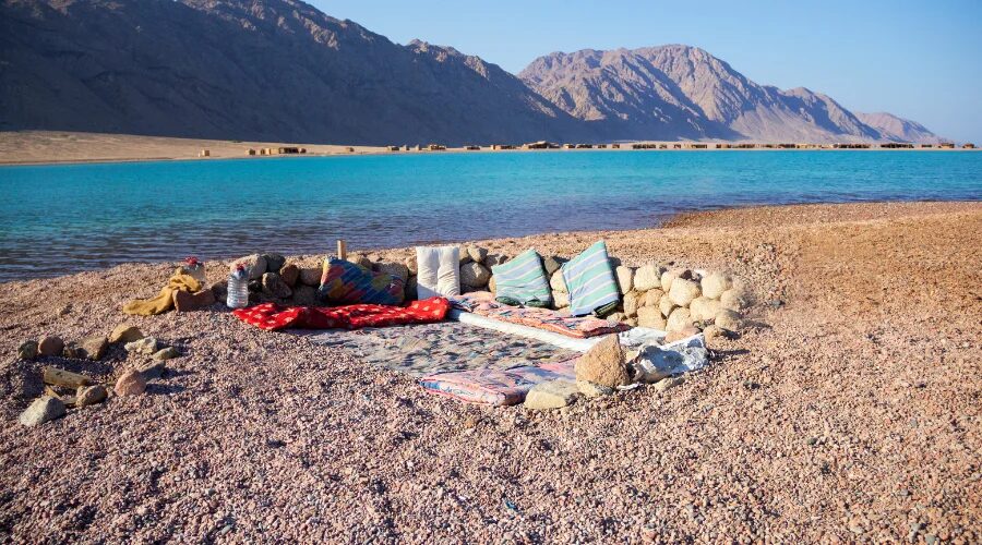 Bedouin-style cushions and rugs on a pebble beach by a turquoise lagoon below the mountains near Dahab