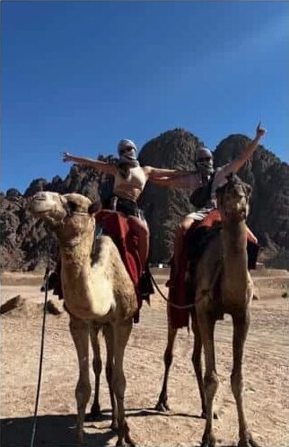 Two riders with arms outstretched on camels in front of dark Sinai mountains