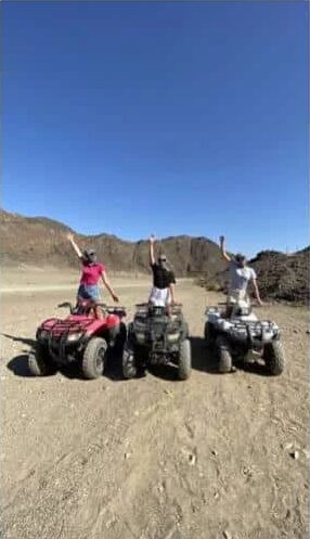Three quad bike riders standing on their quads on a desert track below the Sinai mountains