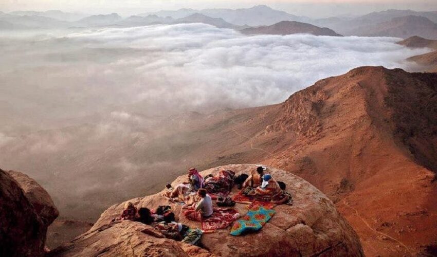Hikers resting on rugs on a rock ledge high above a sea of clouds on Mount Sinai