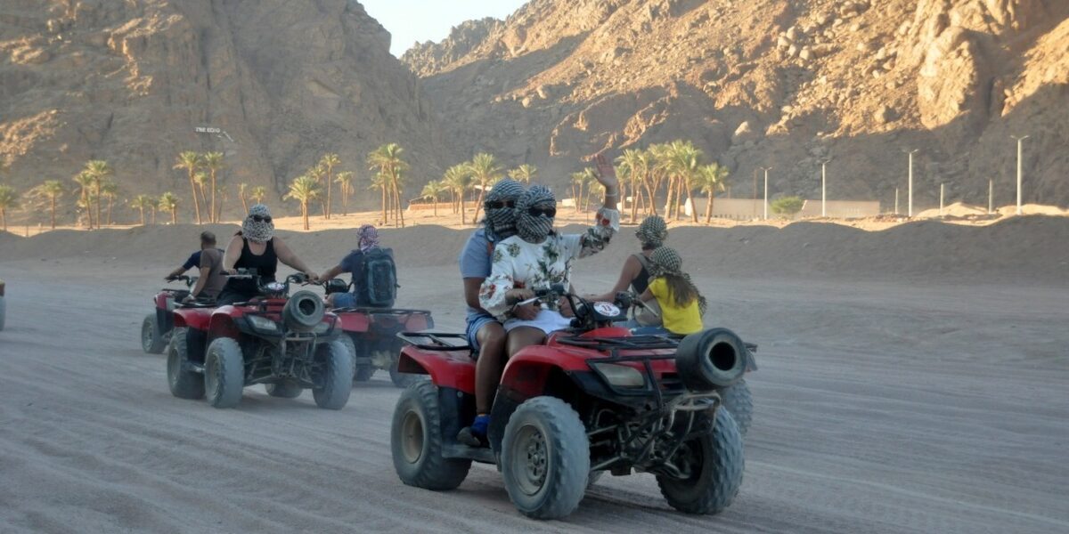 Small group riding red quad bikes through a desert valley lined with palm trees, one rider waving