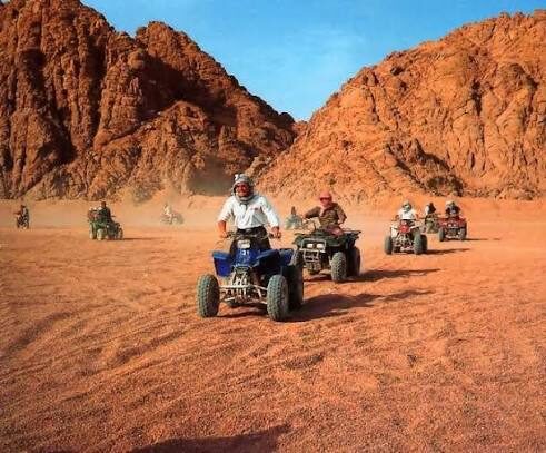 Line of quad bikes riding across orange sand beneath red Sinai mountains